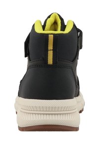 Zapatilla alta de tela negra con forro amarillo, una pestaña en la parte trasera para tirar y una suela blanca con acentos texturizados; presenta un detalle de franja amarilla.