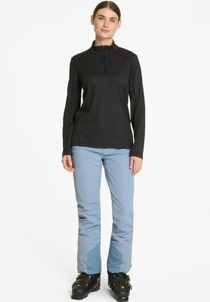 Schwarzes Langarmshirt mit Reißverschluss am Hals und hellblaue Skihosen mit verstärkten Knien. Schwarze Skischuhe mit metallischen Beschlägen.