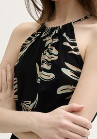 Žena s překříženýma rukama, oblečená v bezrukávovém černém halter topu s béžovými a světle modrými abstraktními vzory na jednobarevném světlem pozadí.