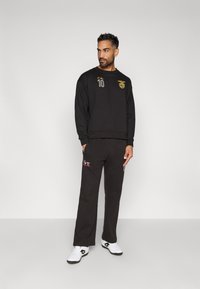 Čierna mikina s kultivovanými zlatými logami a číslom 10, spárovaná s čiernymi nohavicami s červenými akcentmi a bielymi športovými topánkami.