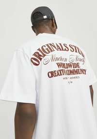 Camiseta de algodón blanca con corte oversize. En la espalda presenta un gran texto marrón: "ORIGINAL STUDIOS" y "COMUNIDAD CREATIVA MUNDIAL", con un diseño curvado.