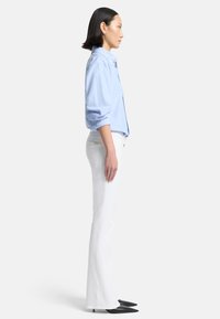 Camicia azzurro chiaro con maniche arrotolate, abbinata a pantaloni bianchi a campana e tacchi a punta neri, vista di lato.