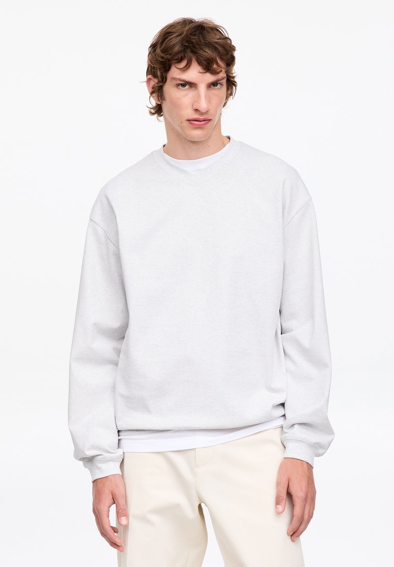 Jeune homme aux cheveux bruns ondulés portant un sweatshirt gris clair uni et un pantalon crème, debout devant un fond blanc, regardant devant lui.