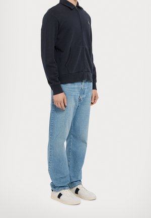 Mies, jolla on musta vetoketjutakki, vaaleansiniset suorat farkut ja valkoiset lenkkarit mustilla raidoilla, seisoo yksiväristä taustaa vasten.