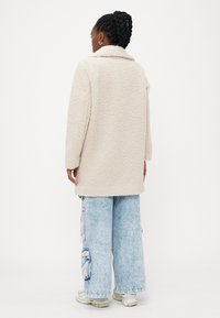 Harmaa beige ylipitkä fleece-takki, jossa on kauluspää, teksturoitu pinta ja sivutaskut, yhdistettynä vaaleansinisiin löysiin farkkuihin.