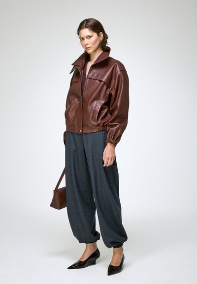 Jeune femme portant une veste en cuir marron, un pantalon ample gris foncé, portant un sac à main marron et des chaussures noires à bout pointu, debout sur fond blanc.