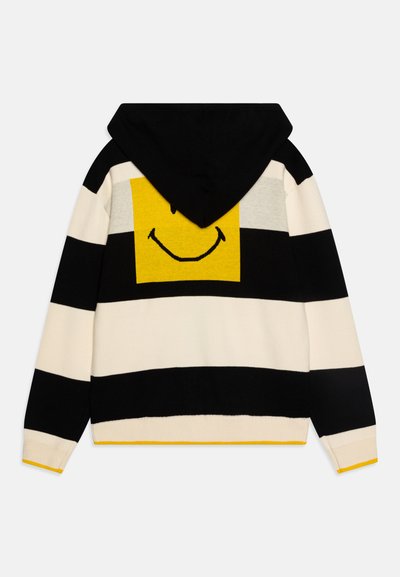 Felpa con cappuccio a righe nei colori nero e crema; presenta un quadrato giallo con una grafica di un sorriso sul retro. Texture morbida a maglia, cappuccio regolabile.