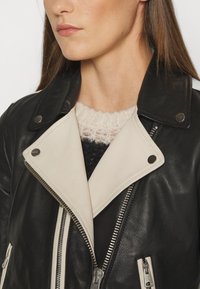 Chaqueta de cuero negra y crema con cierre de cremallera diagonal, solapas anchas y botones a presión. Textura suave con paneles de color contraste.