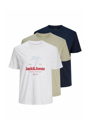 Ung mand med krøllet hår iført en hvid Jack & Jones t-shirt, blå denimshorts, beige sko og hvide sokker, står på en ensfarvet baggrund.