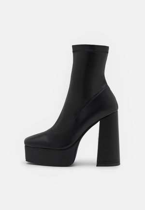 Bottine à plateforme noire avec partie supérieure extensible, talon haut épais et bout arrondi sur fond blanc.