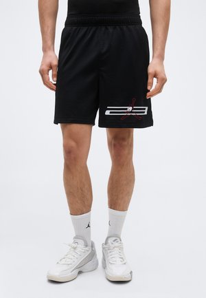 Homme portant un short de basketball noir avec un logo Jumpman blanc et rouge, des chaussettes blanches et des baskets blanches sur un fond uni.