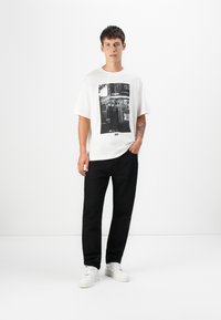 White graphic t-shirt with a black and white photograph print, paired with black pants and white sneakers. Simple, casual design and fit.