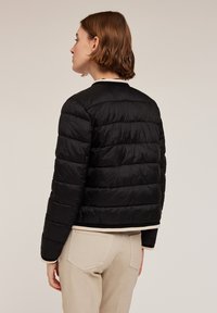 Giacca a vento nera con texture a trapunta orizzontale, colletto e polsini a costine, lunghezza corta. Indossata sopra pantaloni chiari.