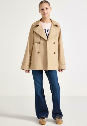 Ung kvinde iført beige dobbeltradet frakke, lyserød sweater med mørkt mønster, blå jeans og beige sneakers mod ensfarvet baggrund.