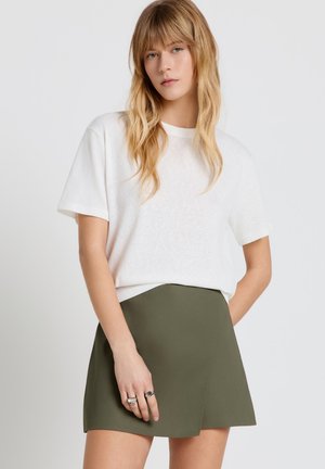 Junge Frau mit langen blonden Haaren, die ein weißes kurzärmeliges Shirt und einen olivgrünen Wickelrock trägt, steht vor einem schlichten Hintergrund.