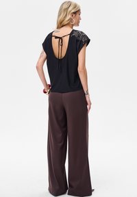 Czarny top z odkrytymi plecami i wiązaniem, ozdobiony detalami na ramionach, zestawiony z szerokimi brązowymi spodniami.
