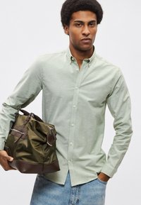 Πράσινο-ανοιχτό πουκάμισο με υφή, συνδυασμένο με τσάντα duffel σε σκούρο πράσινο και καφέ, με φερμουάρ και χερούλια.