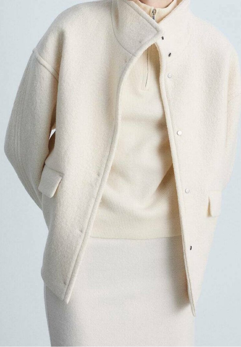 Nainen, joka on pukeutunut vaaleankreemiväriseen villatakkiin raidoitetun vetoketjupuseron ja siihen sopivan vaaleankreemivärisen hameen päälle, seisoo tasaisella taustalla.