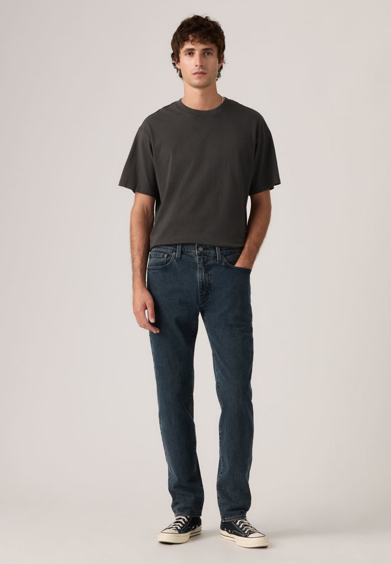 Μπλούζα κοντομάνικη σκούρου γκρι χρώματος συνδυασμένη με μπλε τζιν παντελόνι ίσιας γραμμής και μαύρα αθλητικά παπούτσια, στέκεται μπροστά σε ουδέτερο φόντο.