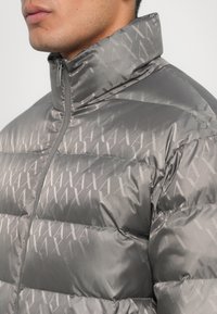 Jachetă puffer gri cu guler înalt, având un finisaj lucios și un model subtil cu logo pe întreaga suprafață; design quilted cu cusături vizibile.