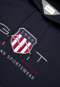 Felpa blu navy con un grande scudo ricamato, strisce rosse e bianche, "GANT 1949" in testo bianco e foderatura interna grigia.