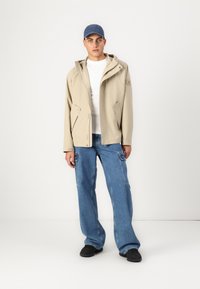 Beige vanntett jakke med hette, hvit t-skjorte, blå denim cargo-bukser og svarte sko, med synlige lommer og logo-detall.