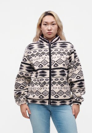 Jeune personne aux longs cheveux blonds portant une veste polaire zippée à motif noir et blanc et un jean bleu clair, debout devant un fond blanc.