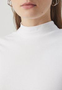 Witte geribbelde katoenen top met hoge neklijn en gladde textuur, met een aansluitend model en een hoge kraag. Zilverkleurige oorringen zichtbaar.