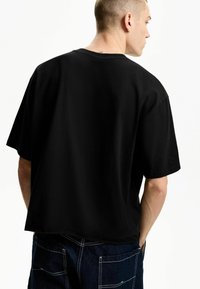 Tricou negru cu mânecă scurtă și guler rotund, cu o croială lejeră. Are o textură netedă și un design minim. Este purtat cu pantaloni din denim de culoare albastru închis.