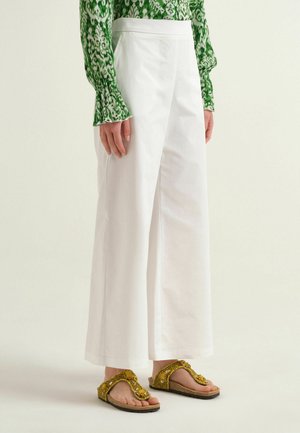 Persona che indossa pantaloni bianchi a gamba larga, una maglietta verde a maniche lunghe con motivo e sandali gialli aperti con perline, in piedi su uno sfondo semplice.