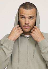 Veste à zip vert clair avec une capuche à cordon, des poignets côtelés et une texture lisse. Le mannequin tient la capuche avec les deux mains.