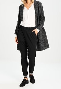 Pantalons de jogging noirs ajustés avec taille élastique et accents rayés, associés à une blouse blanche et un manteau texturé gris. Des baskets noires complètent la tenue.