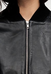 Veste aviateur en cuir noir avec un col côtelé et une fermeture éclair argentée, présentant une texture lisse et des détails de couture minimalistes.