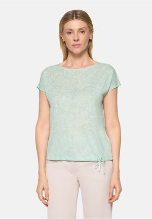 Femme portant un chemisier à manches courtes vert clair à motif floral avec un ourlet à cordon et un pantalon beige, debout devant un fond uni.