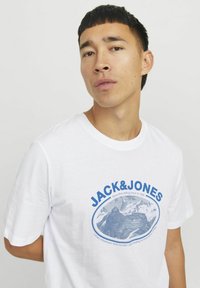 Hvid bomulds T-shirt med et blåt grafisk motiv af bjerge og teksten "JACK&JONES" i fede bogstaver. Rund hals, korte ærmer.