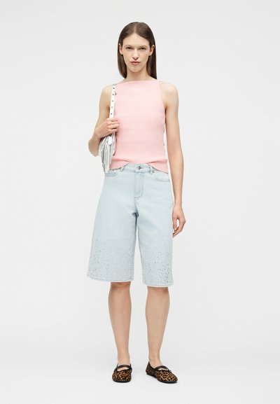 Γυναίκα με ροζ μπλουζάκι χωρίς μανίκια, ανοιχτό μπλε τζιν σορτς μέχρι το γόνατο με διακοσμητικά, μοκασίνια με λεοπάρ σχέδιο και τσάντα ώμου με φούντες.
