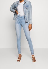 Chaqueta de mezclilla, azul claro, corte clásico con bolsillos frontales. Camiseta gris, jeans ajustados de tiro alto azul claro y sandalias blancas con tiras.