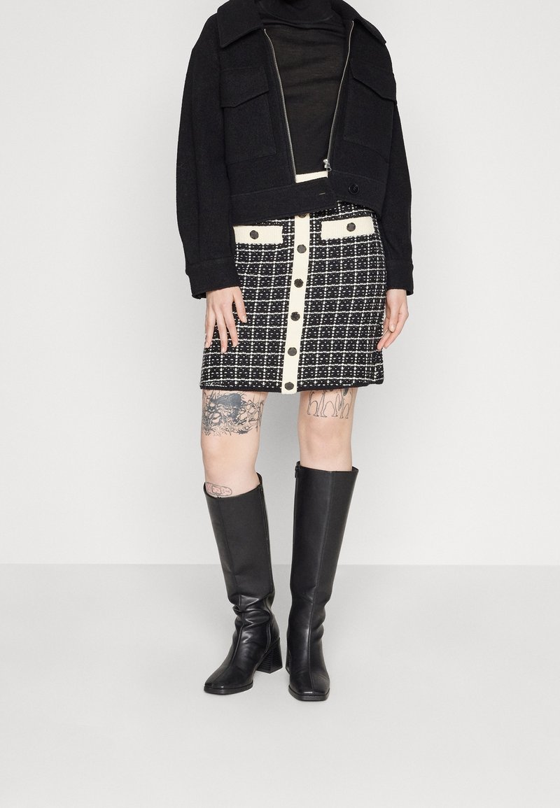 Černá zkrácená bunda s velkými kapsami, v kombinaci s černobílou kostkovanou sukní s knoflíkovými detaily. Černé vysoké boty po kolena.
