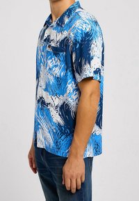 Košile s krátkým rukávem v modrých tónech s vlnkovým vzorem. Vyrobena z lehké látky, má strukturovaný límec a kapsu.