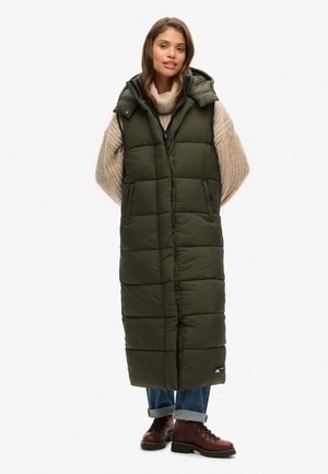 Langer olivgrüner Puffer-Westen mit Kapuze, Frontreißverschluss und zwei seitlichen Taschen. Texturiertes Gewebe mit einem gesteppten Design. Tragen über einem cremefarbenen Pullover.