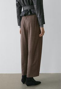 Person iført brune bukser med vide ben, sorte ruskind ankle-støvler og en sort læderjakke med peplumkant, stående på et gråt gulv.