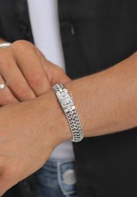 Sidabrinės metalinės grandinėlės apyrankė su sujungtomis grandelėmis ir segtuku, ant kurio išgraviruotas logotipas. Dėvima ant šviesaus odos tono riešo.