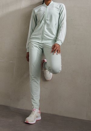 Persoon die een lichtgroen Adidas trainingspak en witte sneakers met roze accenten draagt, staand tegen een simpele muur, één been omhoog.
