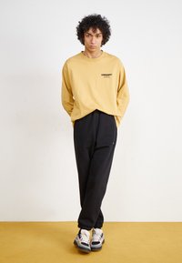 Long-sleeve mustard shirt with black logo, paired with black sweatpants. Footwear features a multicolor design with white and blue accents.