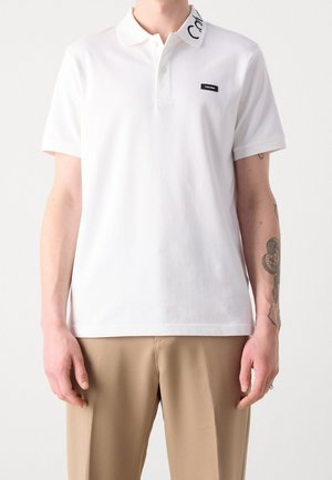 T-shirt polo blanc à manches courtes, design minimaliste, avec un logo noir sur le col et une petite étiquette noire sur la poitrine.