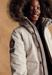 Jachetă puffer bej cu glugă căptușită cu blană, închidere cu fermoar și două buzunare laterale. Include un patch cu logo pe mâneca stângă.
