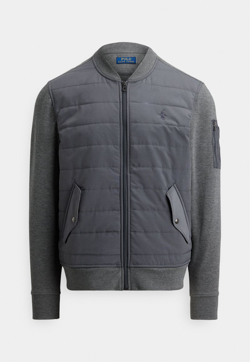 Veste bomber grise avec devant matelassé, manches en maille grise et fermeture éclair. Comprend des poches latérales et un logo sur la poitrine.