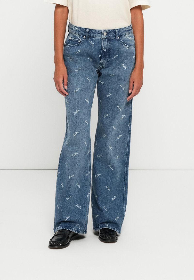 Siniset leveät farkut, joissa on toistuva tekstikuvio, viisi taskua ja klassinen napitus. Yhdistetty mustiin kenkiin.