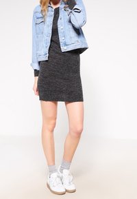 Giacca di jeans azzurro chiaro sopra un abito di maglia grigio scuro, abbinata a sneakers bianche e calzini grigi. Design semplice, forma aderente.