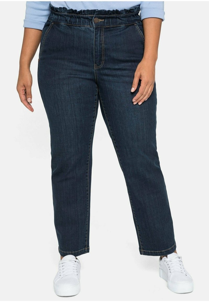 Jean en denim foncé avec une taille froncée, fermeture à bouton à l'avant, poches latérales et coupe droite. Associé à des baskets blanches.